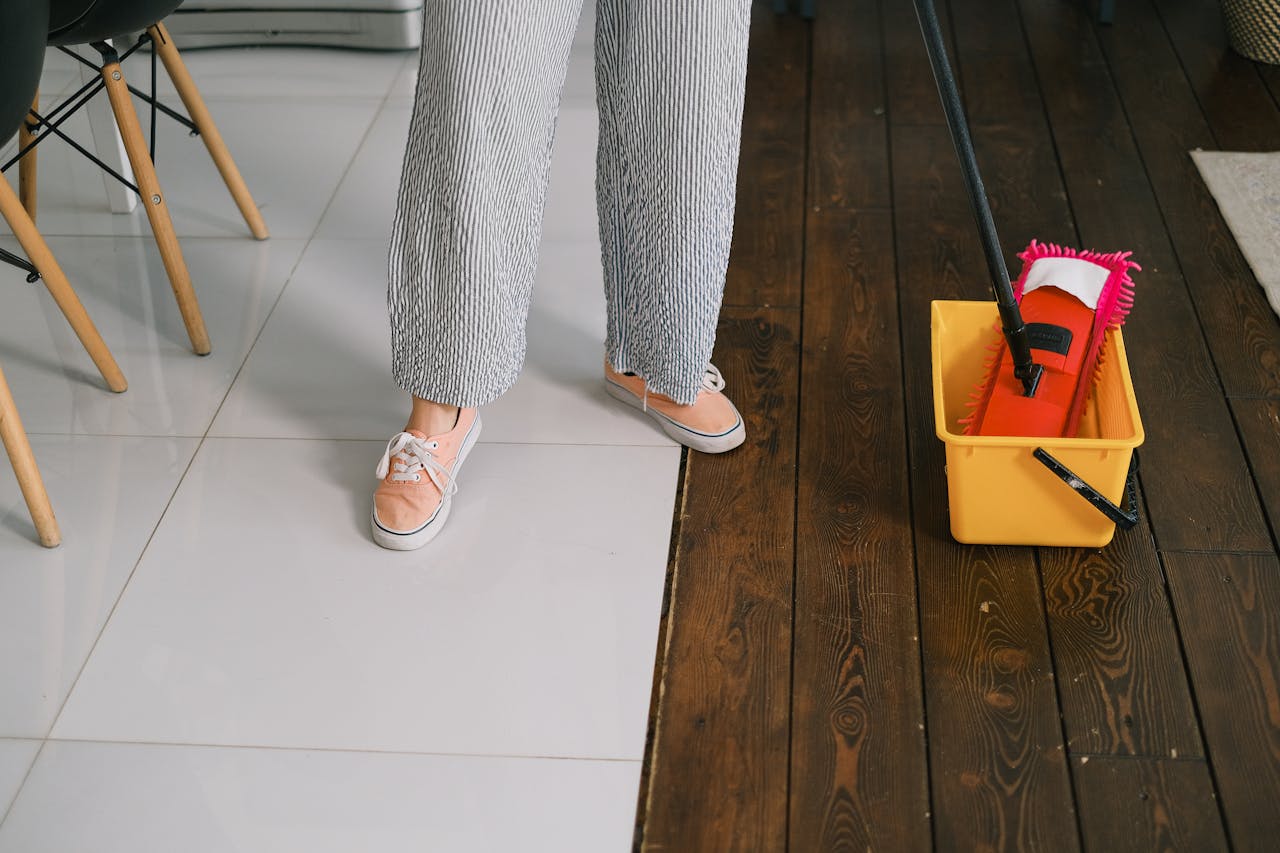 gallery-6 Crop anonymous woman in gumshoes and pants dipping mop into bucket with water on parquet in house