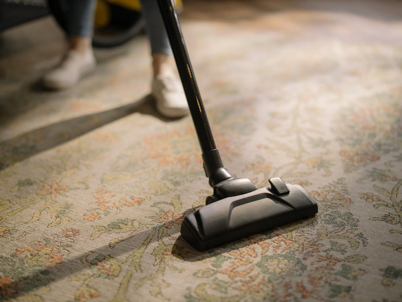 hero-img-02 Close-up of a vacuum cleaner on a patterned carpet in a sunlit room, capturing a moment of household cleaning.